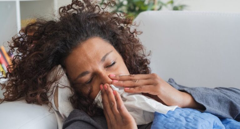 Woman sitting on a sofa blowing her nose into a tissue while feeling unwell, illustrating how to fight back against winter viruses during cold and flu season