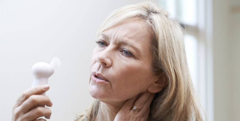 A middle-aged woman holding a small handheld fan and touching her neck, showing discomfort from hot flushes and night sweats.