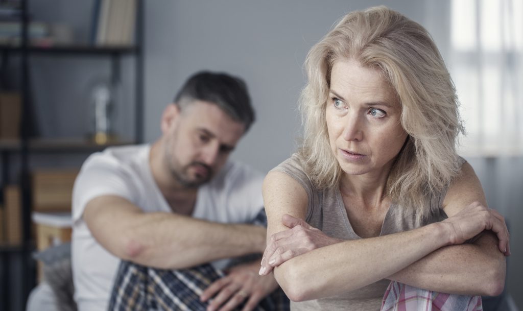 Woman sitting with arms wrapped around herself looking distressed while a man sits in the background, reflecting relationship strain and emotional pain