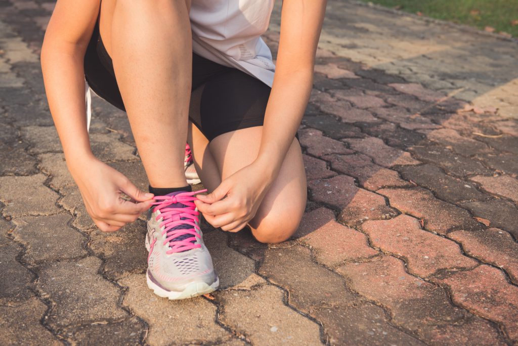 Woman kneeling on a paved path tying her running shoes before exercise, reflecting the importance of movement and strength to support your frame and overall health