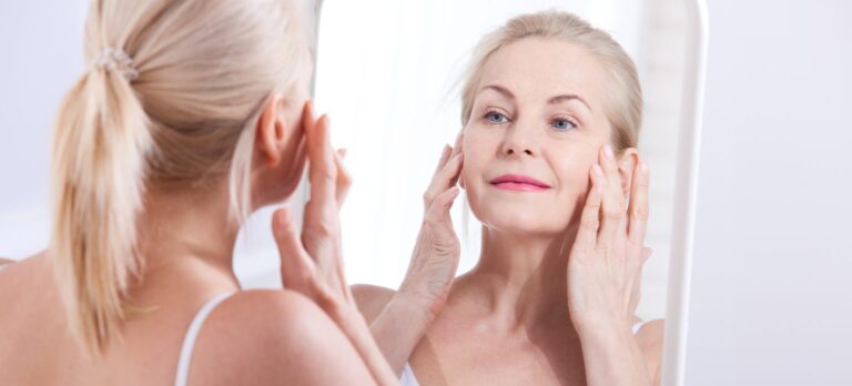 Woman looking in the mirror and gently touching her face, relating to the use of oestrogen cream for facial skin concerns.