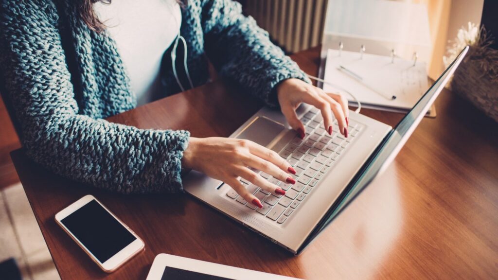 Woman typing on a laptop at a desk with a mobile phone beside her, representing online advocacy and calls to demand action on perimenopause and menopause care.