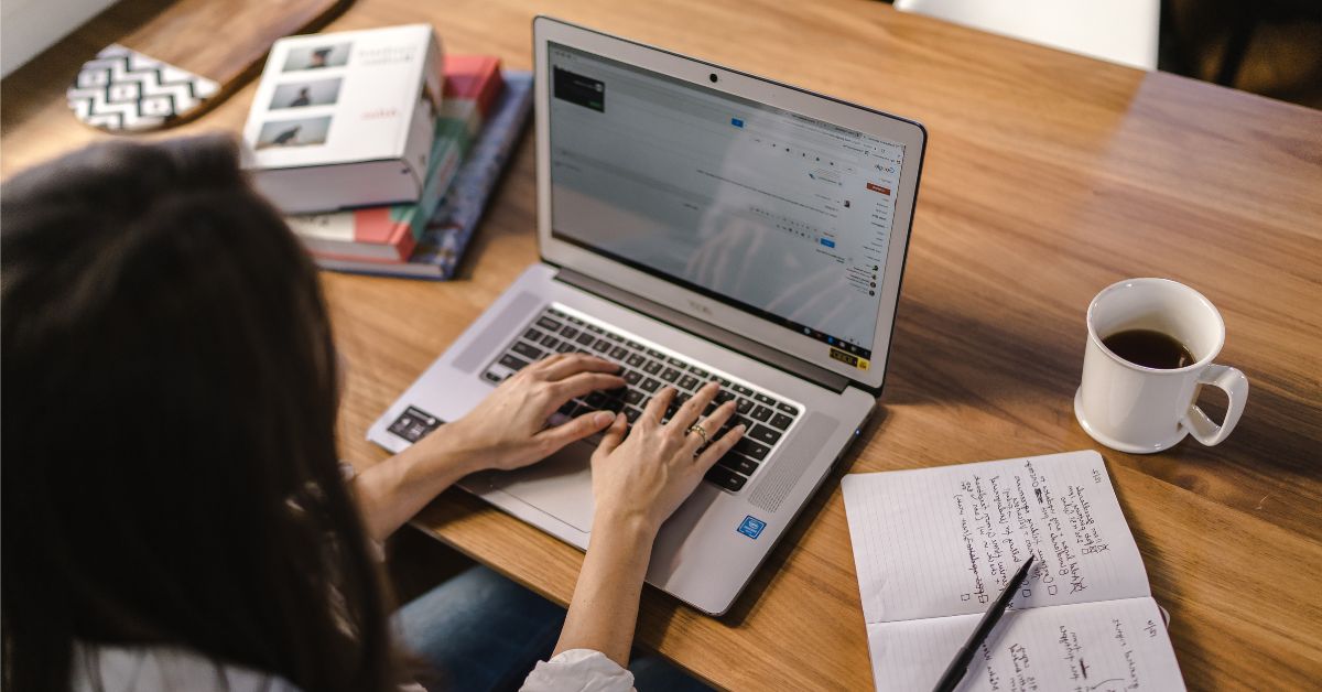Woman typing on a laptop at a desk with notes and a coffee nearby, representing making a submission to the Senate Menopause Inquiry.
