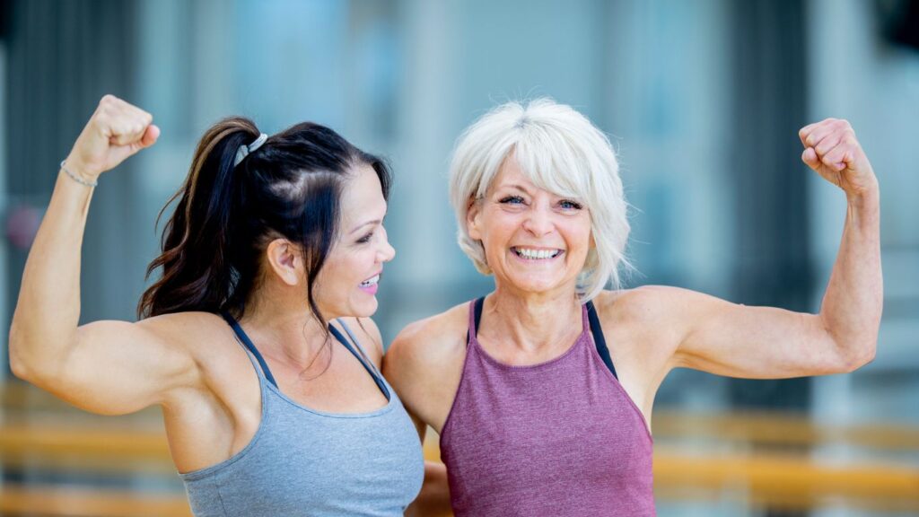 Two women in activewear smiling and flexing their arm muscles, representing strength and fitness after menopause.
