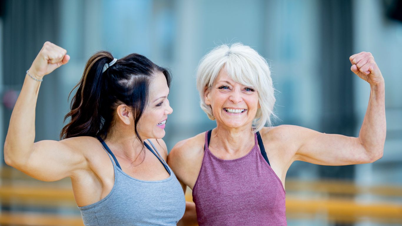Two women in activewear smiling and flexing their arm muscles, representing strength and fitness after menopause.