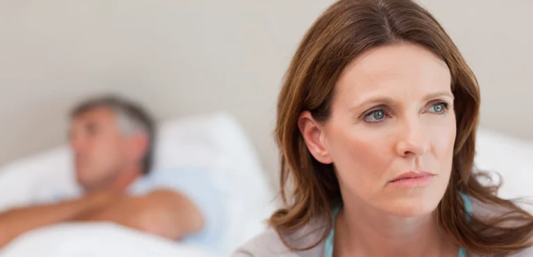 A woman sitting on the edge of a bed looking concerned while her partner lies in the background, illustrating the emotional and relationship impact of vaginal dryness.