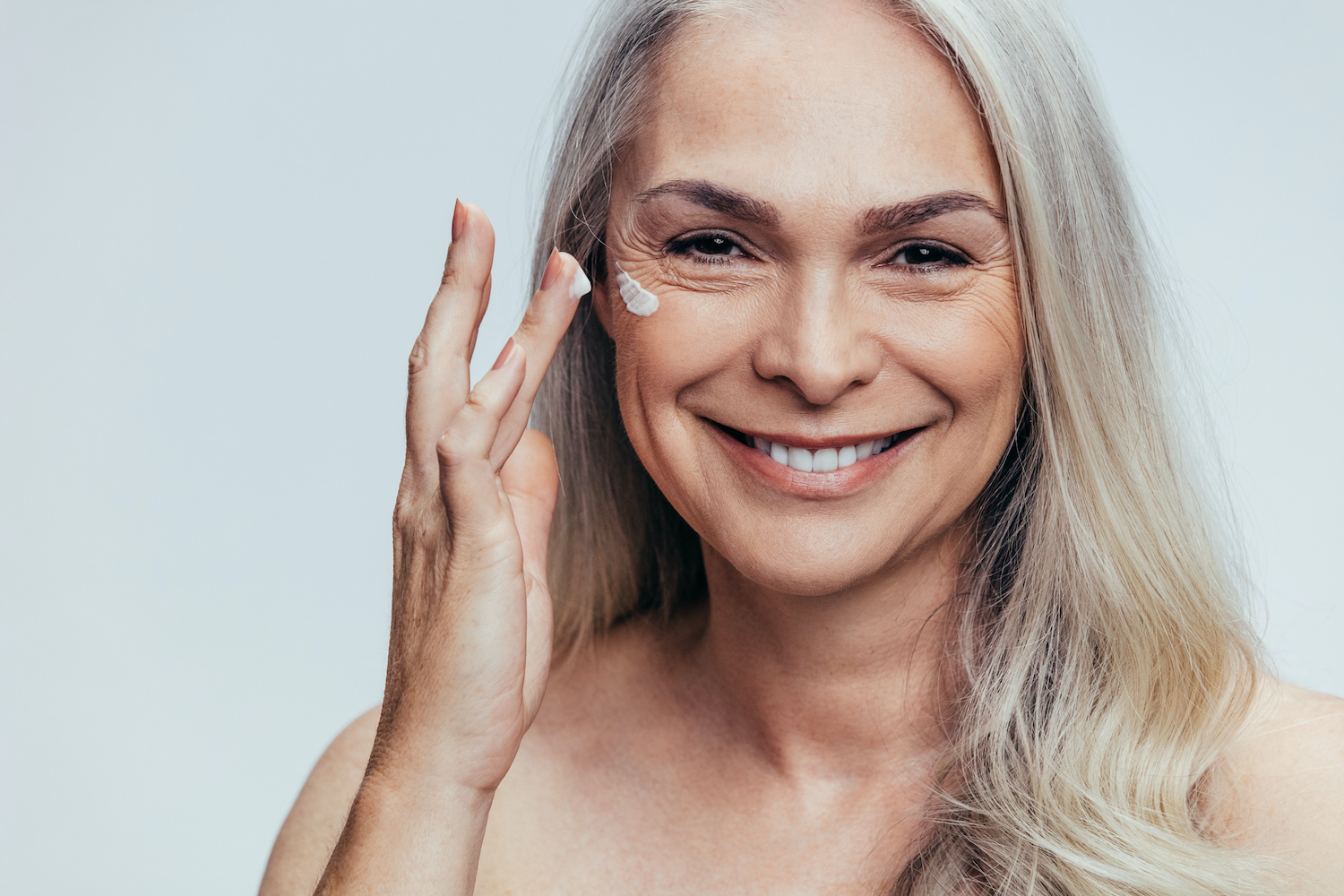 Smiling mature woman applying face cream to her skin, illustrating skincare tips for improving skin at menopause.