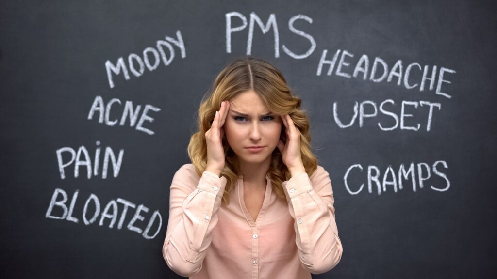 Woman holding her temples with words like “PMS”, “headache”, “cramps” and “moody” written on a blackboard behind her, illustrating hormonal changes during menopause.