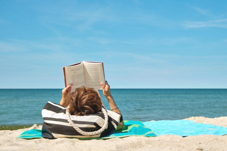 Woman lying on a beach towel reading a book by the sea, representing a relaxing holiday cultural binge guide.