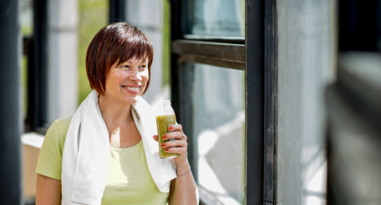 Smiling middle-aged woman holding a fresh juice with a towel over her shoulder, representing healthy diet and exercise after perimenopause.