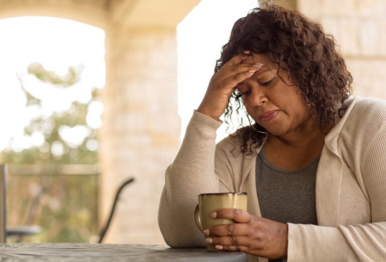 Middle-aged woman sitting at a table holding a mug with her hand on her forehead, illustrating delayed help-seeking for perimenopausal or menopausal symptoms.