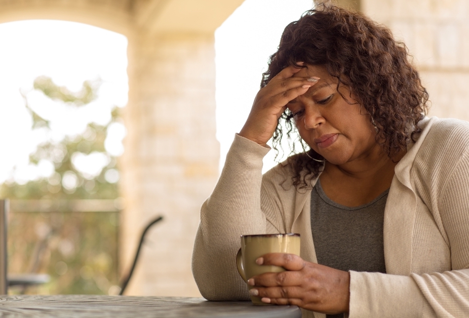 Middle-aged woman sitting at a table holding a mug with her hand on her forehead, illustrating delayed help-seeking for perimenopausal or menopausal symptoms.