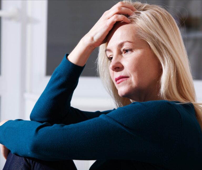 Middle-aged woman sitting on a sofa with her hand on her forehead, looking concerned, illustrating anxiety and heart palpitations around menopause.