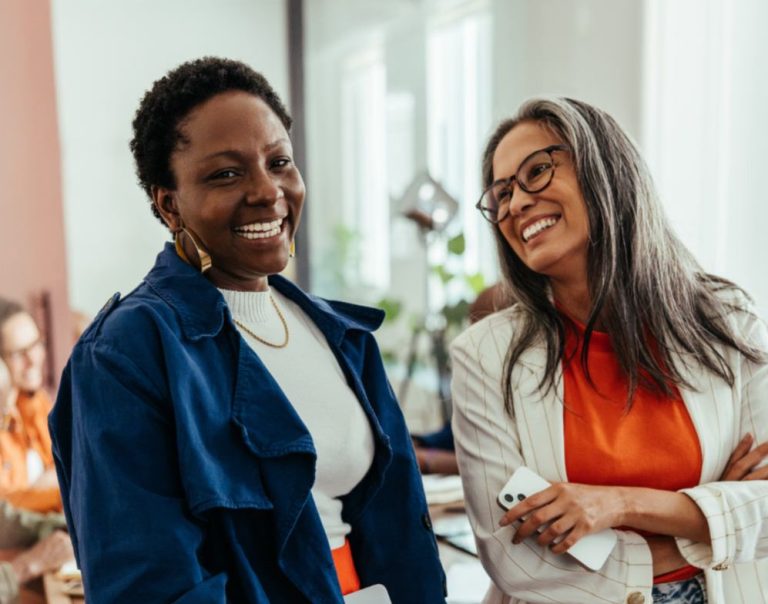 Two female colleagues smiling and chatting in an office, representing open conversations and menopause awareness in the workplace.